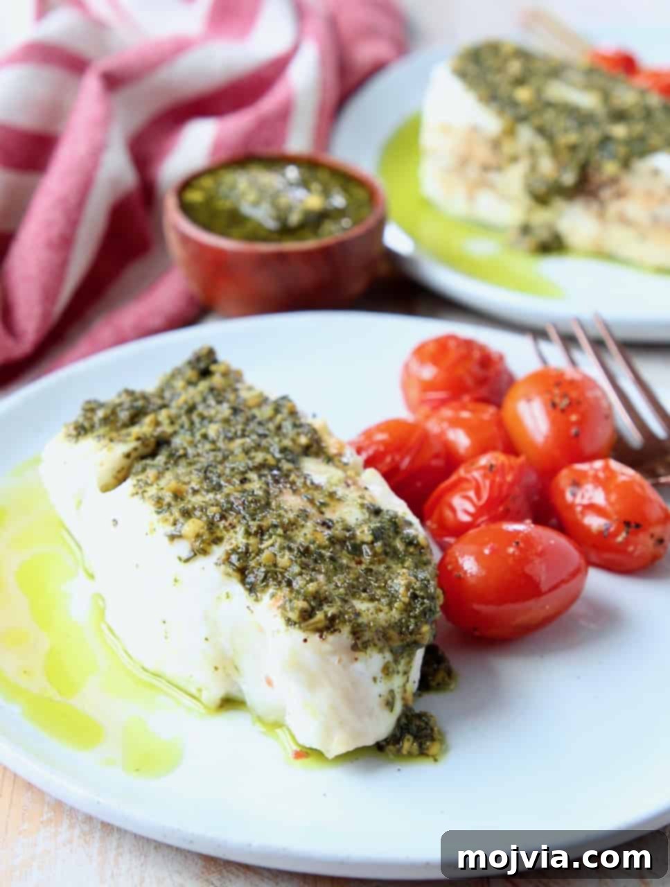 Pesto-Crusted Chilean Sea Bass 3 Close-up of perfectly cooked Chilean sea bass fillet, glistening with basil pesto, served on a white plate alongside vibrant roasted cherry tomatoes and a fresh basil sprig. The fish's tender, flaky texture is visible.