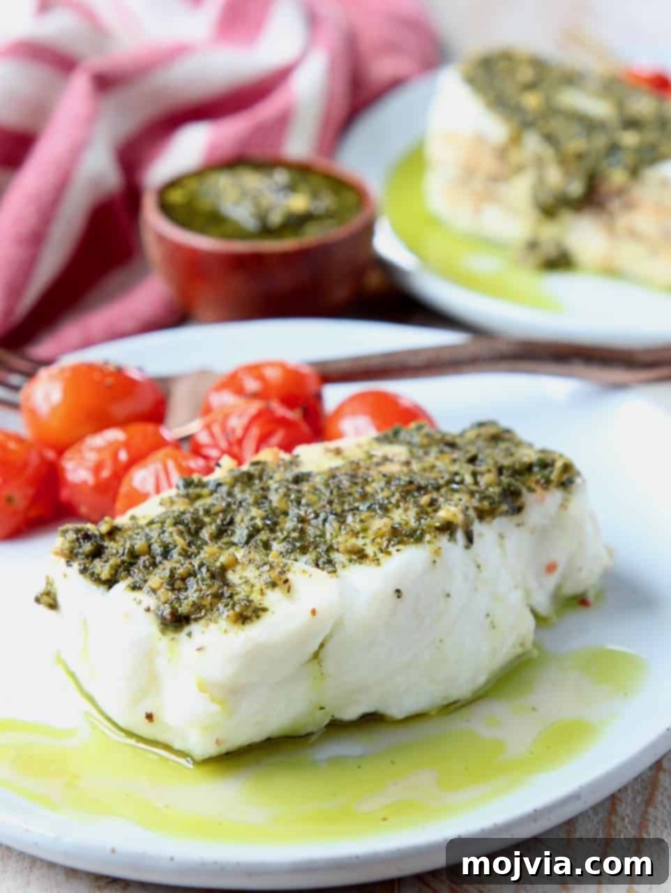 Pesto-Crusted Chilean Sea Bass 4 Two fillets of Chilean sea bass, expertly cooked and generously topped with green basil pesto, presented on a minimalist white plate. The fish appears moist and flaky, ready to be enjoyed.