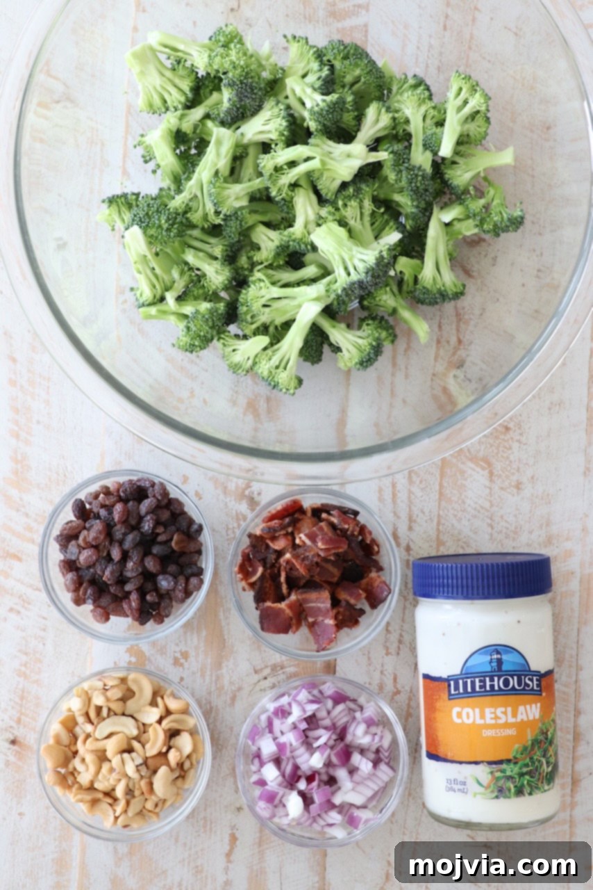 Arrangement of fresh ingredients for making broccoli salad: broccoli florets, red onion, raisins, cashews, and bacon bits on a white wooden board.