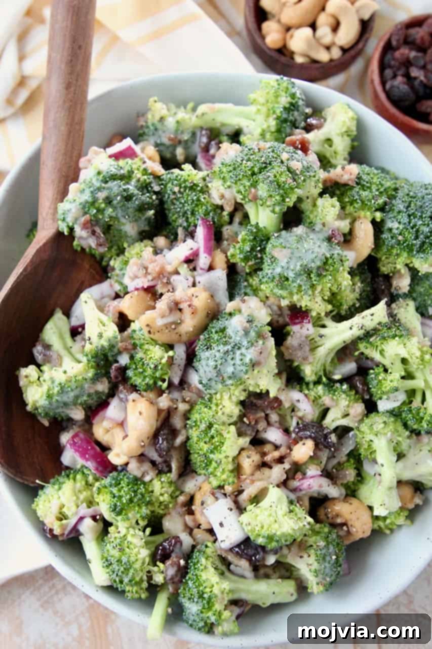 Broccoli salad garnished with a few extra cashews and bacon bits, served in a rustic white bowl with a large wooden serving spoon, ready to be enjoyed.