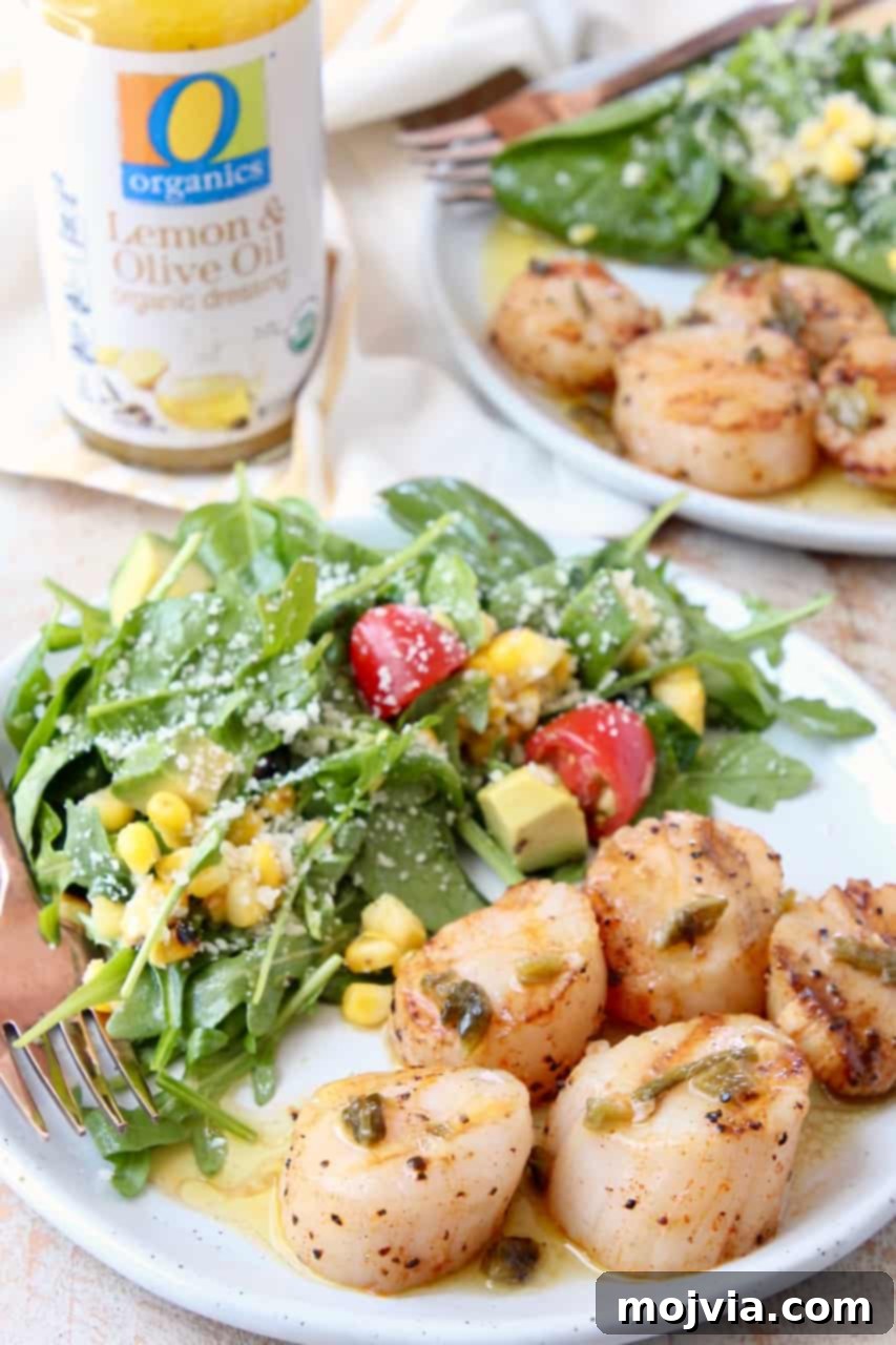 Grilled Scallops with Zesty Jalapeño-Lemon Butter 4 A close-up of grilled scallops on a white plate, glistening with butter sauce, beside a fresh green arugula salad. The scallops show beautiful grill marks.