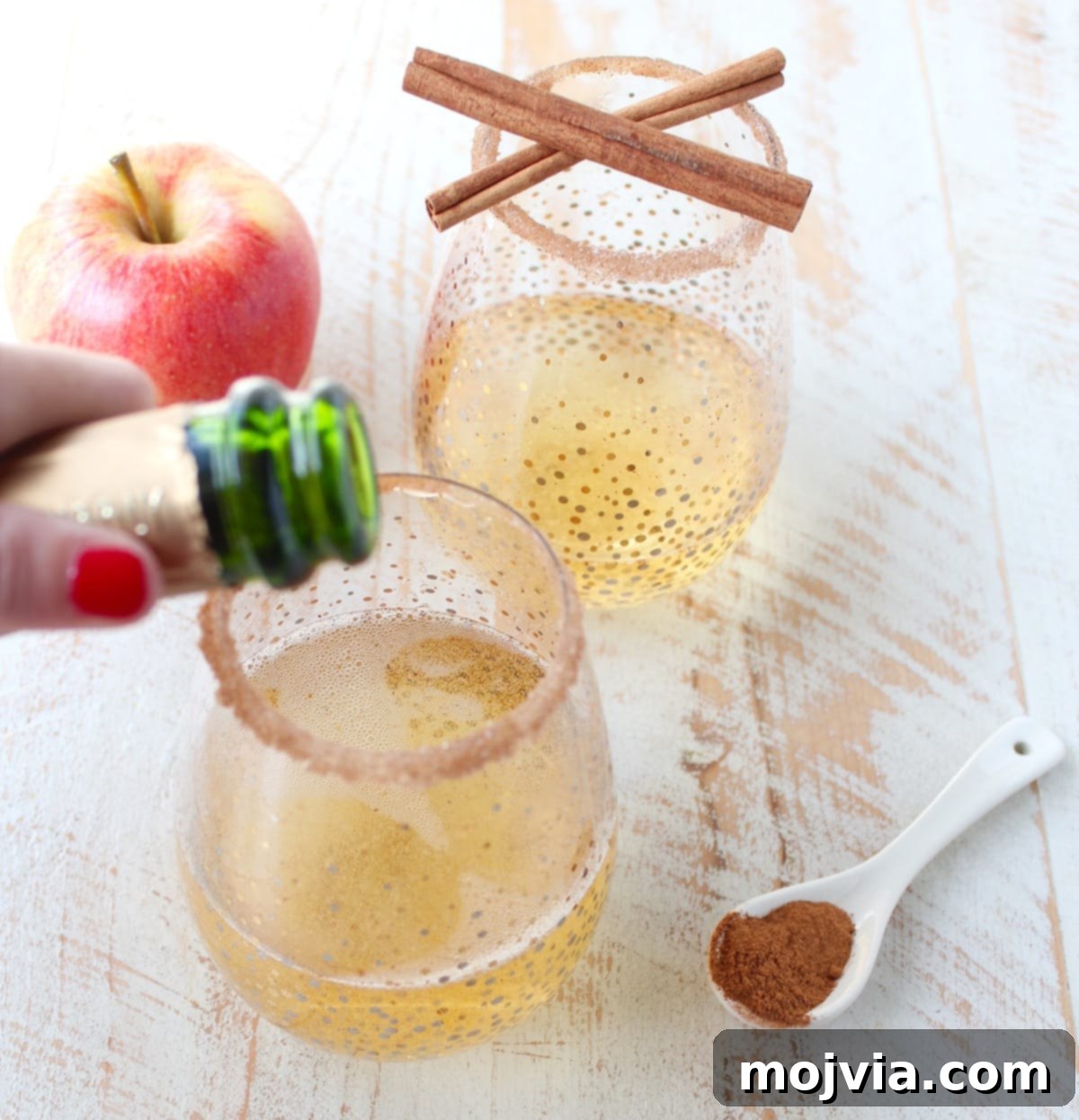 Crisp Apple Champagne Fizz 3 Rich apple cider cocktail being topped with champagne, garnished with apple slices and cinnamon sticks, highlighting its festive appeal and rich color.