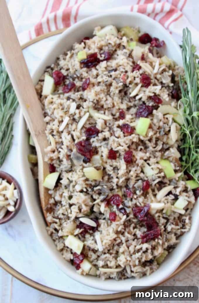 Jeweled Wild Rice Pilaf with Apples and Cranberries 2 Overhead image of Wild Rice Pilaf in a serving dish with a wooden serving spoon, garnished with fresh herbs.
