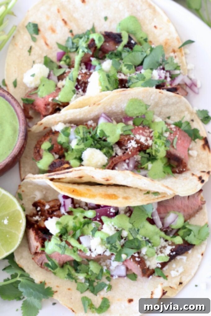 Three perfectly assembled steak tacos on a plate, garnished with fresh cilantro, creamy sauce, and crumbled cheese.