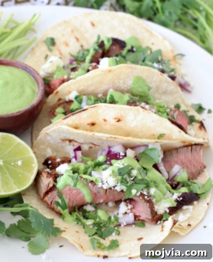 Two steak tacos on a rustic plate, garnished with fresh cilantro, a drizzle of creamy cilantro sauce, and a lime wedge.