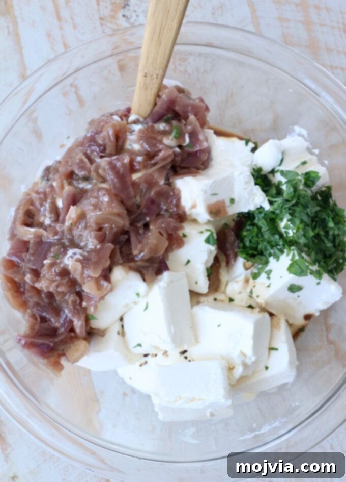 All the French onion dip ingredients are mixed in a clear glass bowl with a spatula, showcasing the creamy texture before chilling.