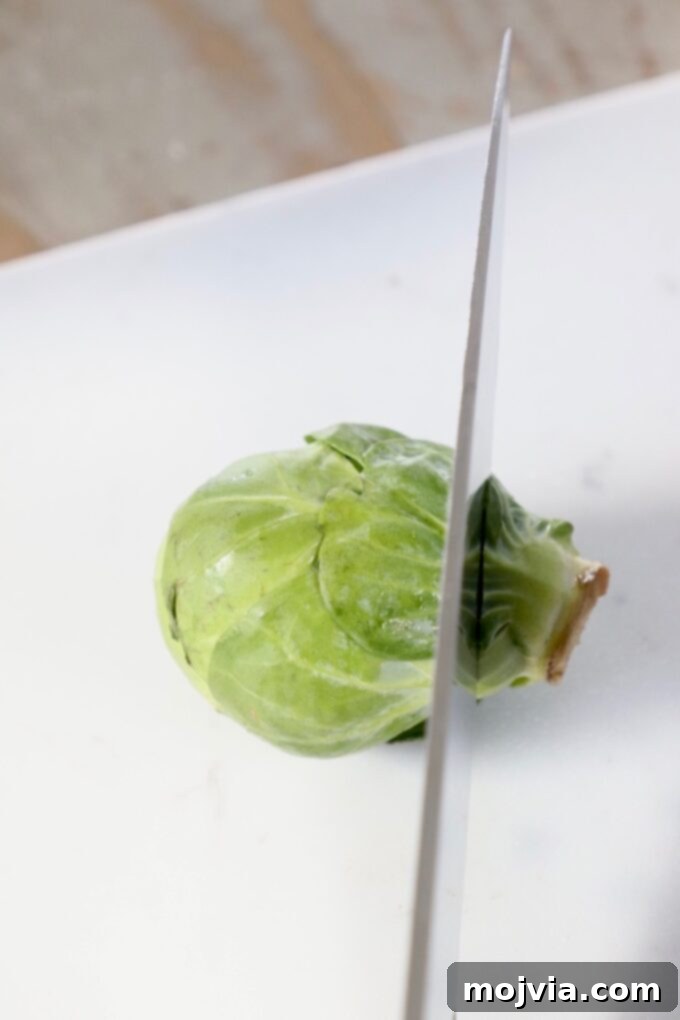Shredding Brussels Sprouts Made Simple 3 A sharp chef's knife beginning to slice into a fresh Brussels sprout on a wooden cutting board, illustrating the initial step of preparation.