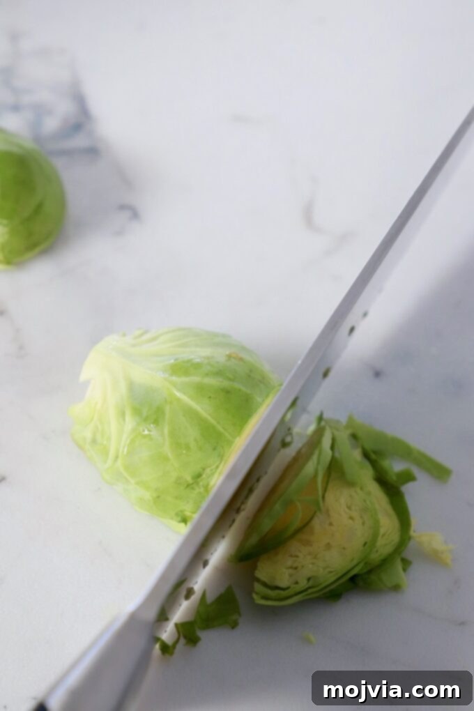 Shredding Brussels Sprouts Made Simple 6 A close-up view of a knife thinly slicing a halved Brussels sprout on a cutting board, demonstrating the shredding technique.