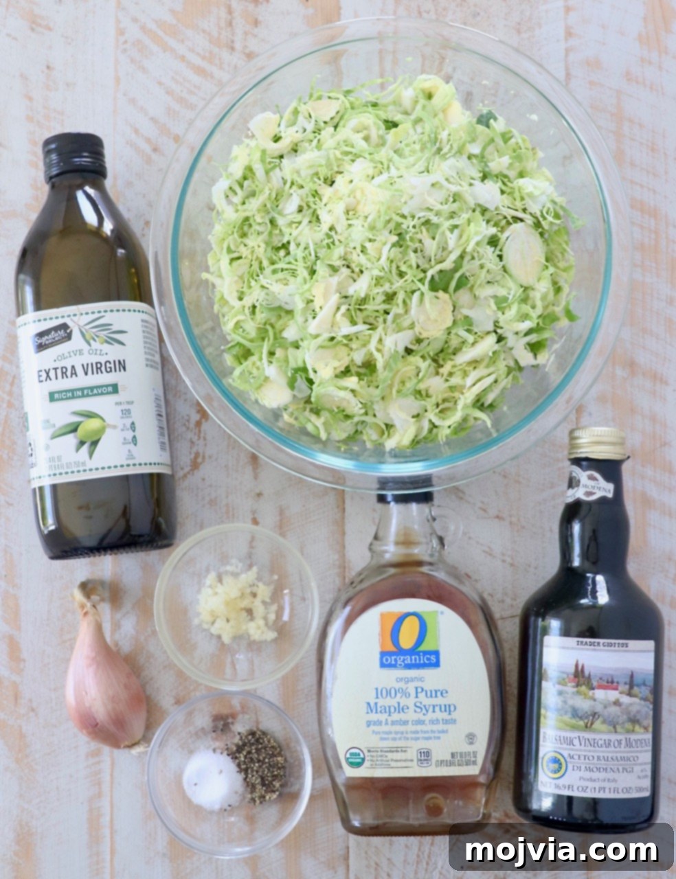 A beautiful flat lay of fresh ingredients for sauteed shaved brussels sprouts including olive oil, fresh brussels sprouts, shallots, garlic, balsamic vinegar, and maple syrup, artfully arranged on a white wooden board.