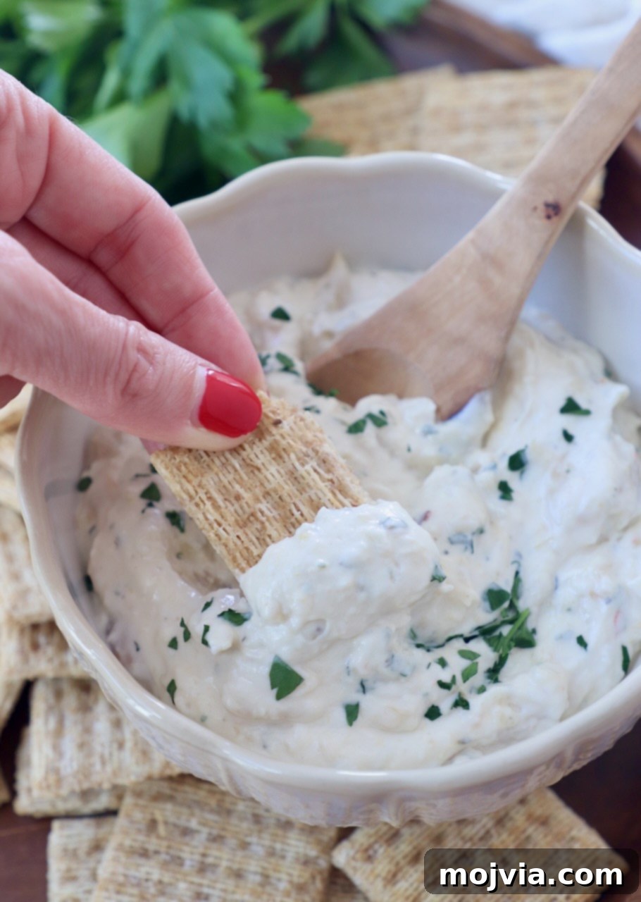 Sweet Roasted Garlic Spread 2 Cracker being dipped into a bowl of creamy roasted garlic dip, next to a wooden spoon.