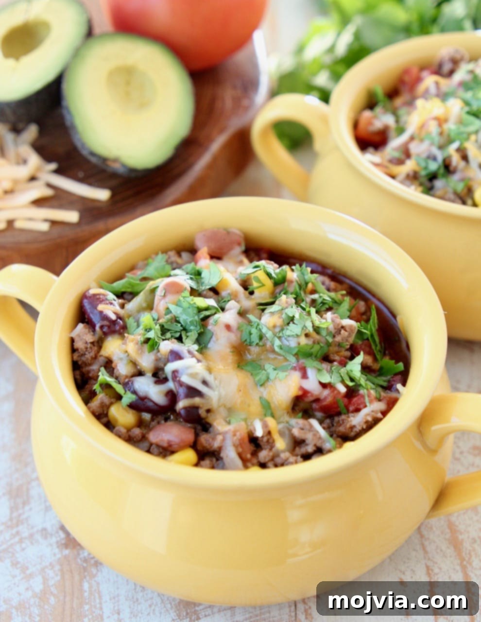 Taco soup in a yellow crock, garnished generously with shredded cheddar cheese and fresh cilantro, served with a gold spoon.