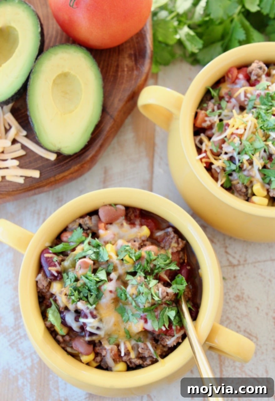 Two yellow crocks of taco soup, garnished with cheddar cheese, fresh cilantro, and diced avocado, served with a gold spoon, next to a red crock pot.