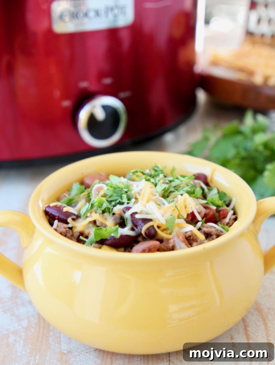 Crockpot taco soup in a yellow bowl with cheddar cheese and fresh cilantro, next to a red crock pot. 