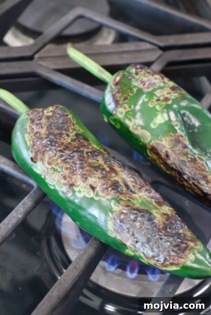 Smoky Roasted Poblanos 8 Roasted poblano peppers on a gas stovetop, with skins visibly charred and bubbling, indicating they are cooked.