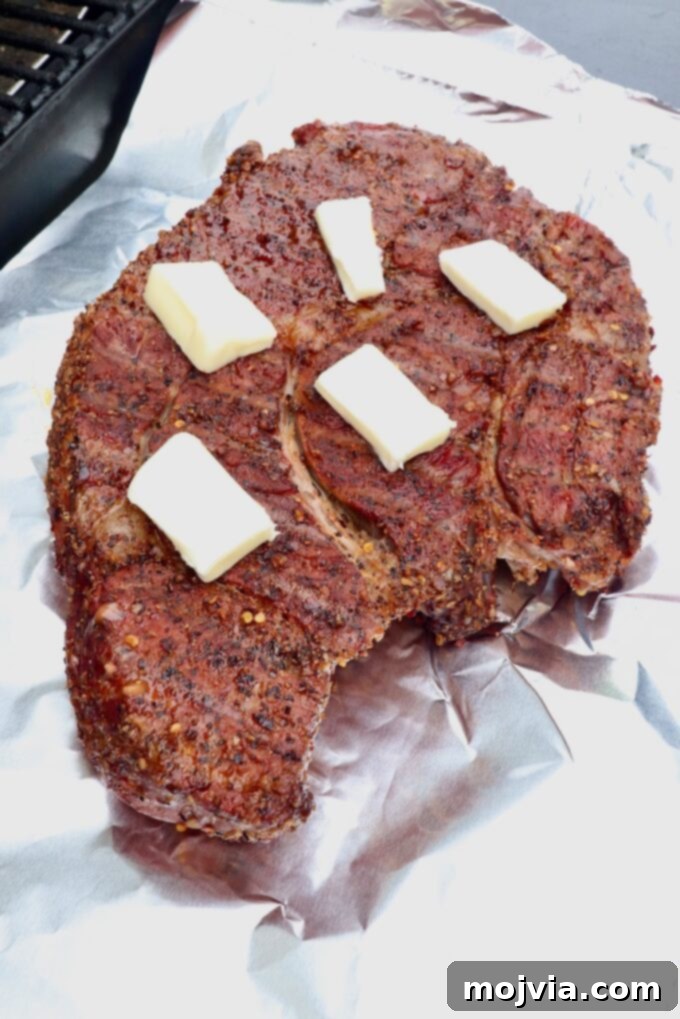 Smoked Beef Chuck Roast Perfection 6 A seasoned smoked chuck roast, partially cooked, with pats of butter melting on its surface, placed on a sheet of aluminum foil before wrapping.