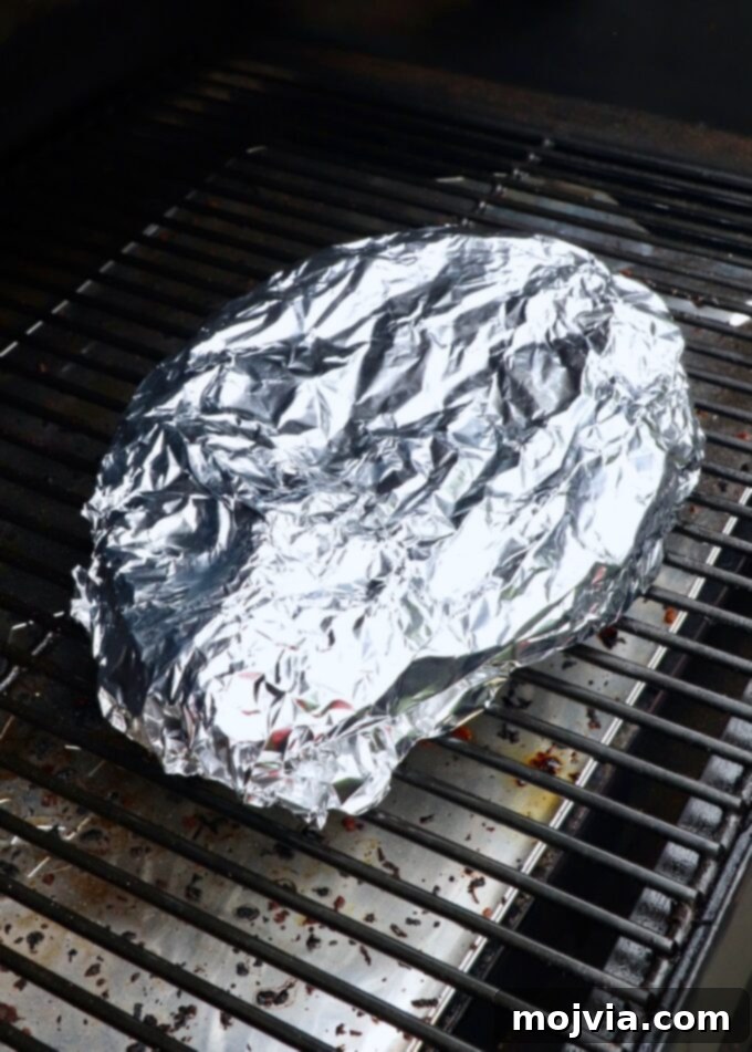 Chuck roast tightly wrapped in foil, sitting on the smoker grates, demonstrating the Texas crutch method.
