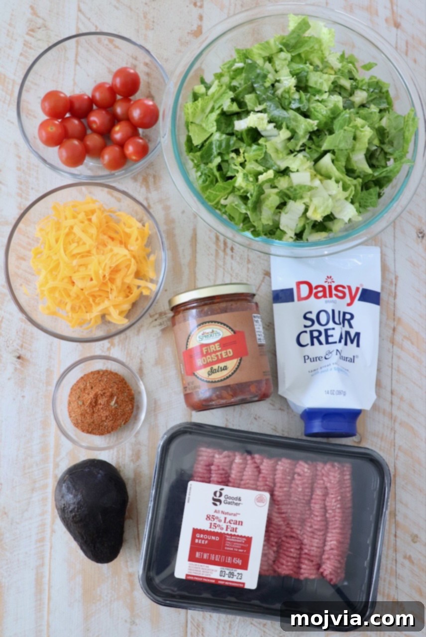 Hearty Beef Taco Salad 3 All fresh ingredients for a homemade taco salad laid out on a white wooden cutting board, including ground beef, lettuce, cheese, tomatoes, avocado, salsa, and sour cream.