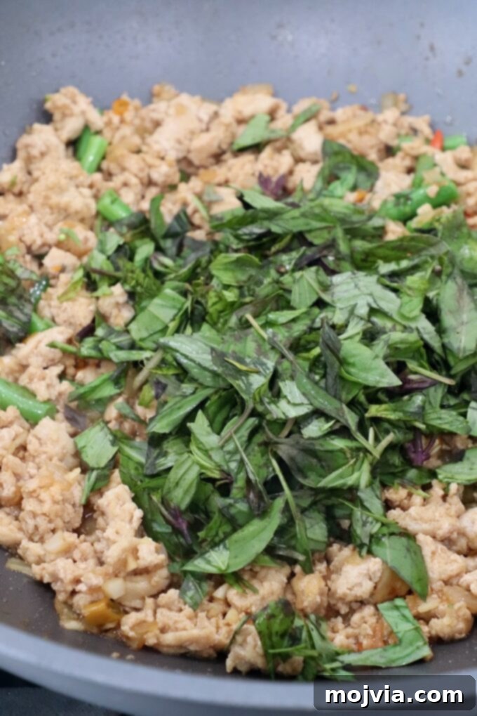 Spicy Thai Basil Chicken Stir-fry 7 Fresh Thai basil leaves being added to the large pan with cooked ground chicken and vegetables, the final step in preparing Thai Basil Chicken.