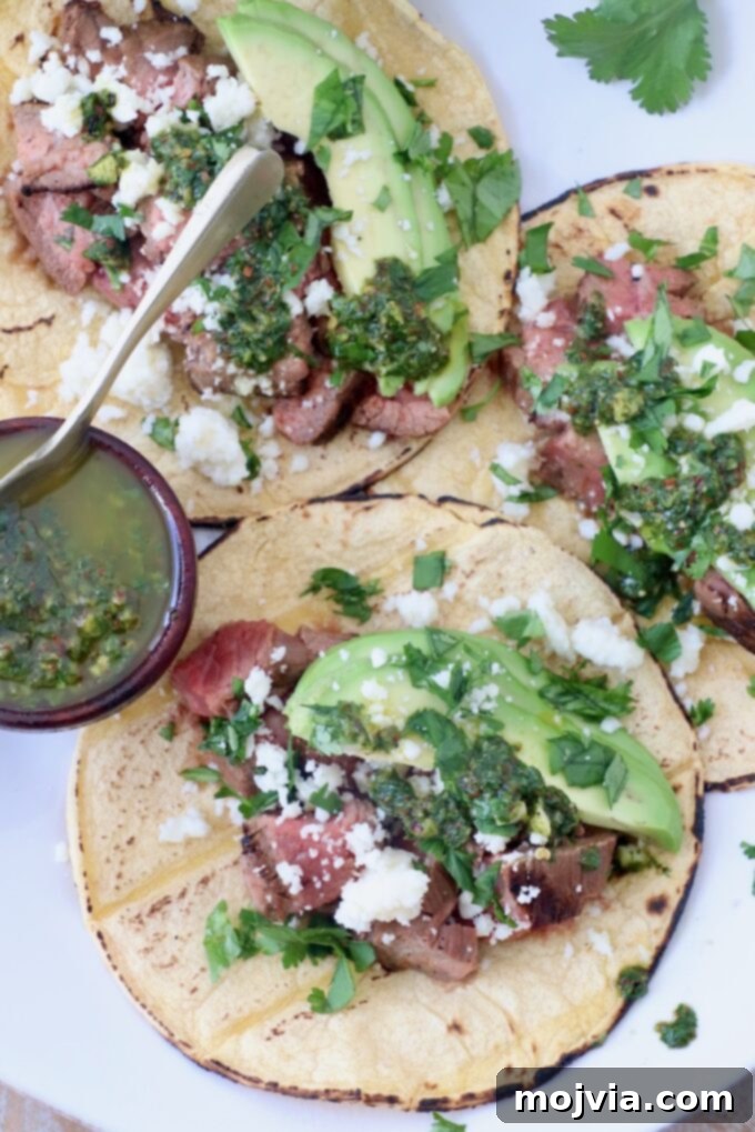 Sizzling Flank Steak Tacos 11 three tacos on white plate with a small bowl of chimichurri sauce on the side