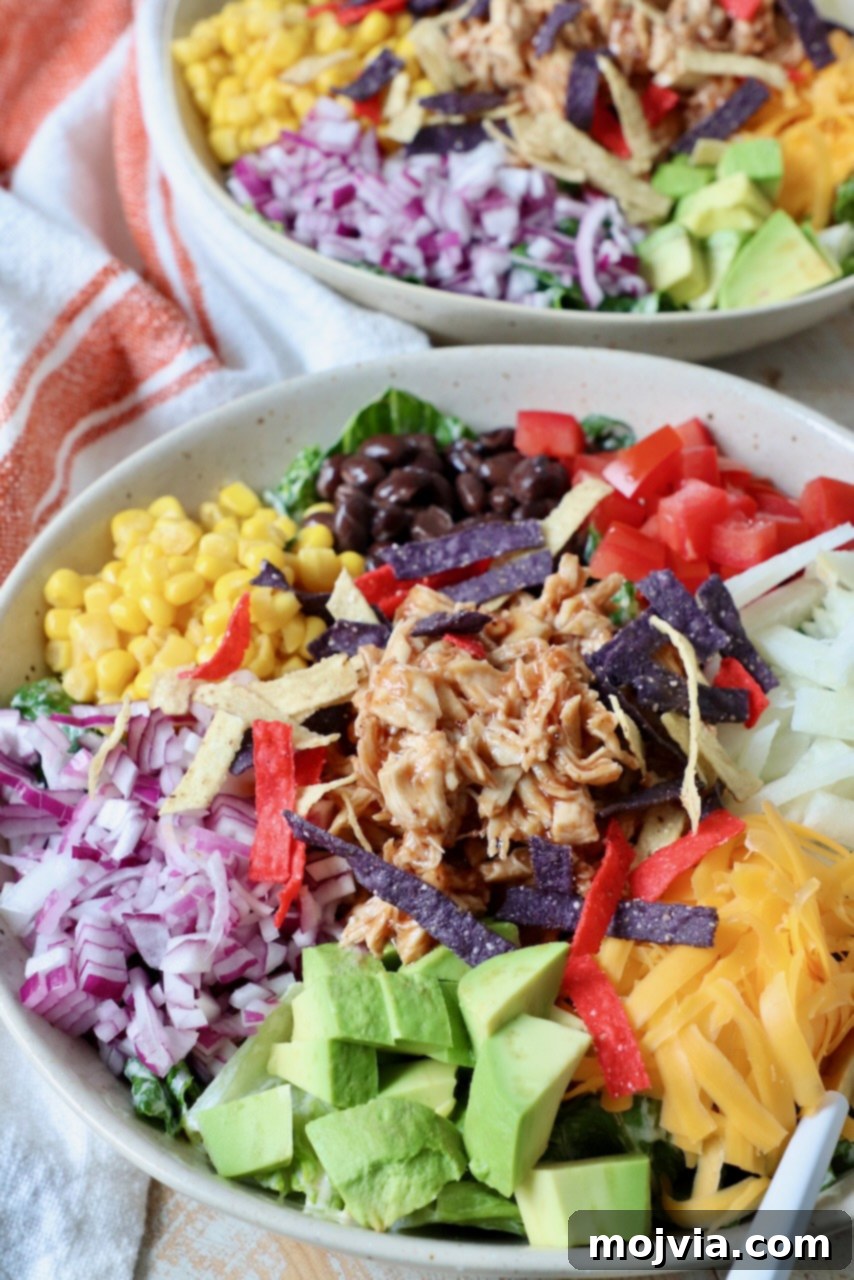 A bowl of BBQ Chicken Salad, showing the layered ingredients: diced onion, avocado, tomatoes, corn, black beans, and shredded cheddar cheese beneath the BBQ chicken.