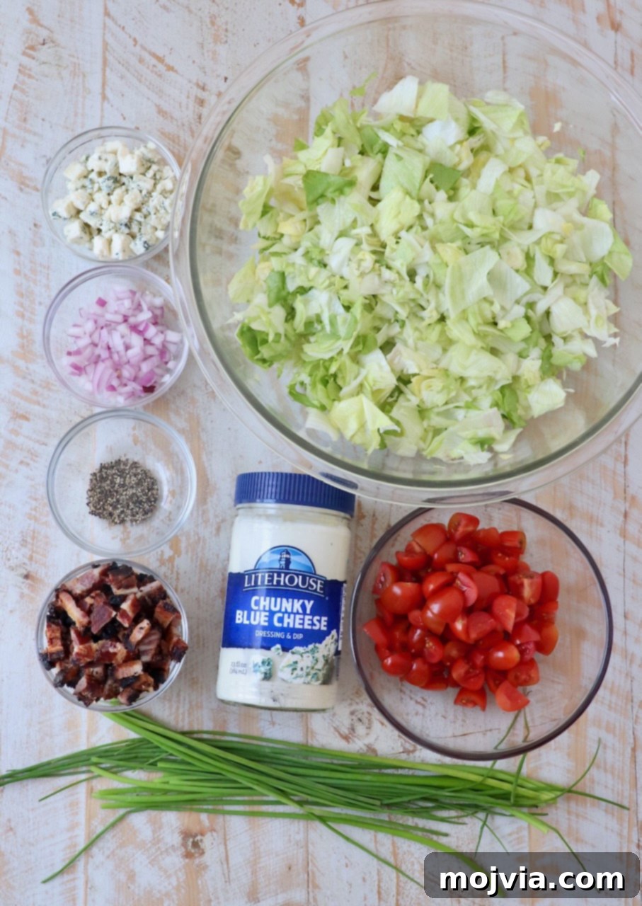 Tangy Blue Cheese Greens 3 Fresh ingredients for chopped blue cheese salad laid out on a white wooden board, including iceberg lettuce, cherry tomatoes, blue cheese crumbles, bacon, red onion, and chives.