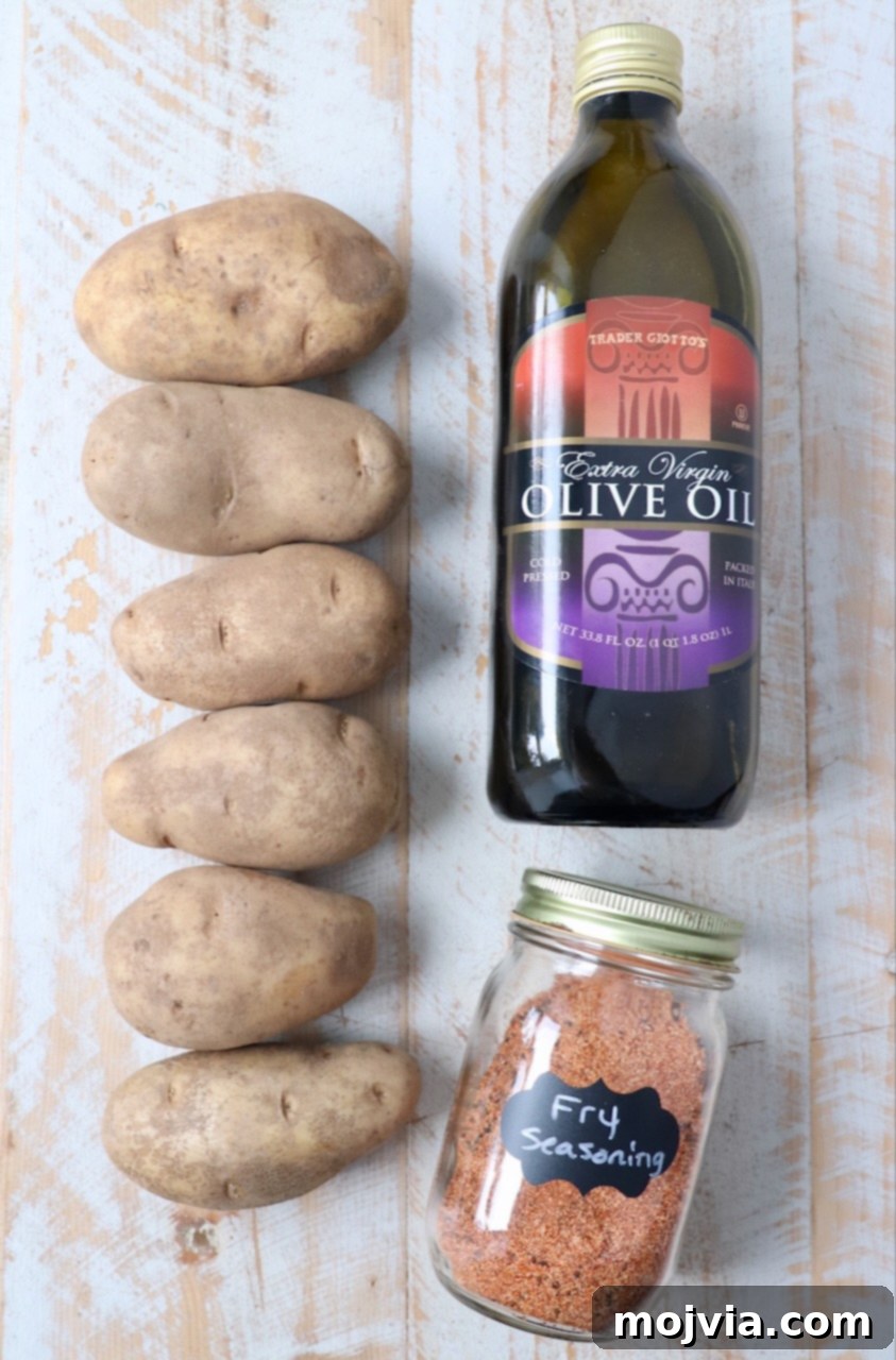 Crispy Air Fryer Fries 3 Ingredients for homemade air fryer french fries laid out on a white wooden board, including russet potatoes, olive oil, and fry seasoning.