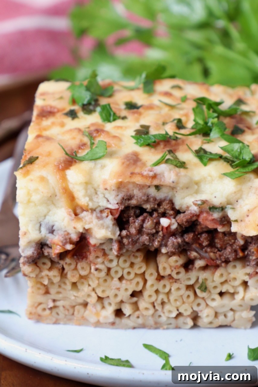 Creamy Greek Comfort Bake 2 A perfectly portioned slice of cooked Pastitsio on a white plate, garnished with fresh chopped parsley.