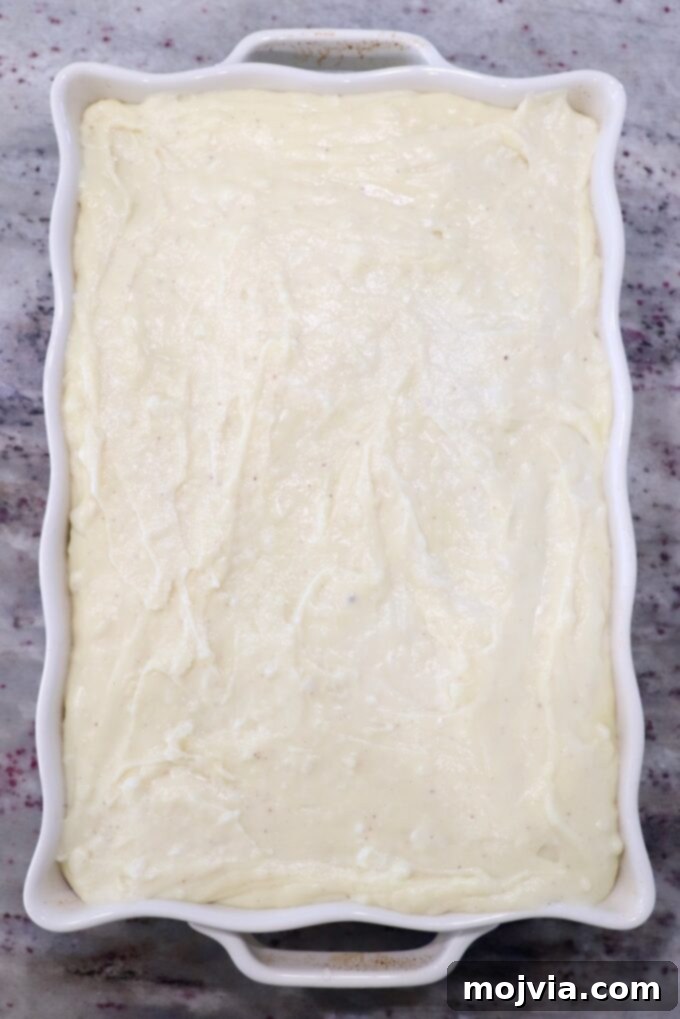 Creamy Greek Comfort Bake 13 The thick, creamy béchamel sauce being poured smoothly and evenly over the meat sauce layer in the casserole dish.