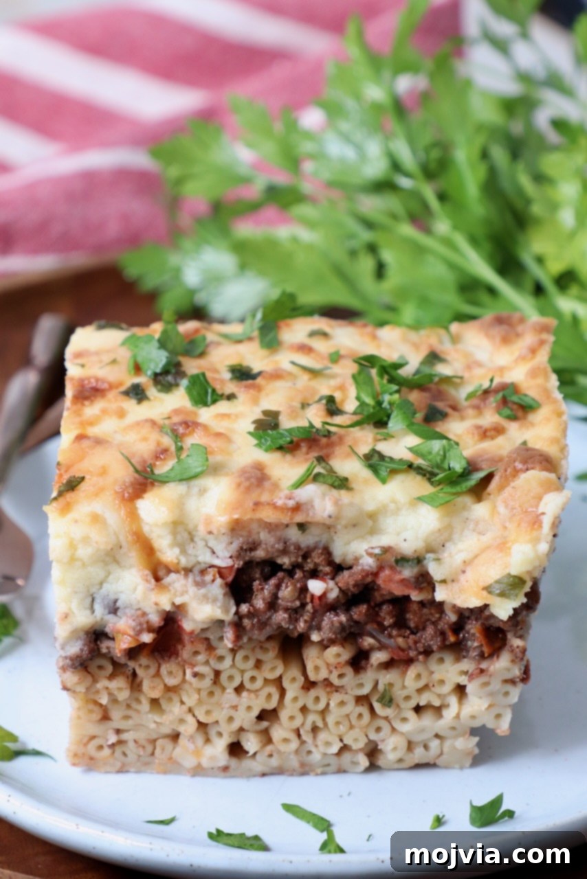 Creamy Greek Comfort Bake 16 A beautiful, individual slice of Pastitsio on a white plate, topped with fresh parsley, highlighting its distinct layers.