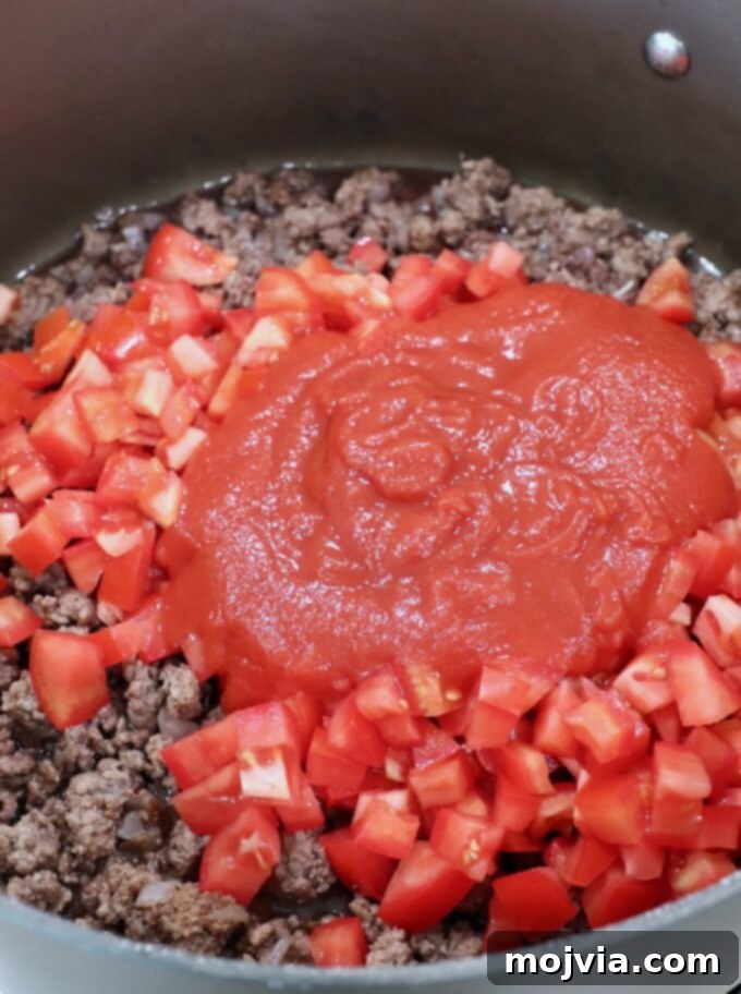 Creamy Greek Comfort Bake 7 Cooked ground beef simmering with diced Roma tomatoes and rich tomato sauce in a large skillet, ready for more ingredients.