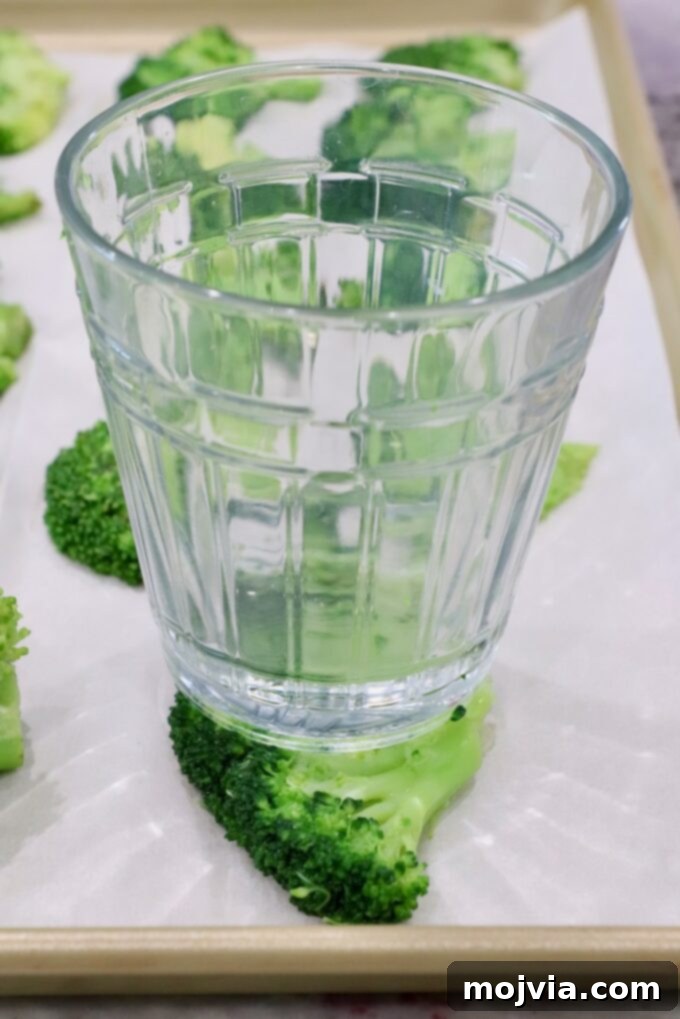 Crispy Smashed Broccoli 7 A glass cup pressing down on a steamed broccoli floret on a parchment-lined baking sheet to smash it flat