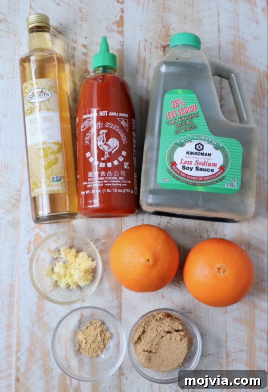 Zesty Orange Glaze 3 Ingredients for making homemade Chinese orange sauce laid out on a white wooden board, including fresh orange, soy sauce, sriracha, garlic, ginger, brown sugar, and white wine vinegar.