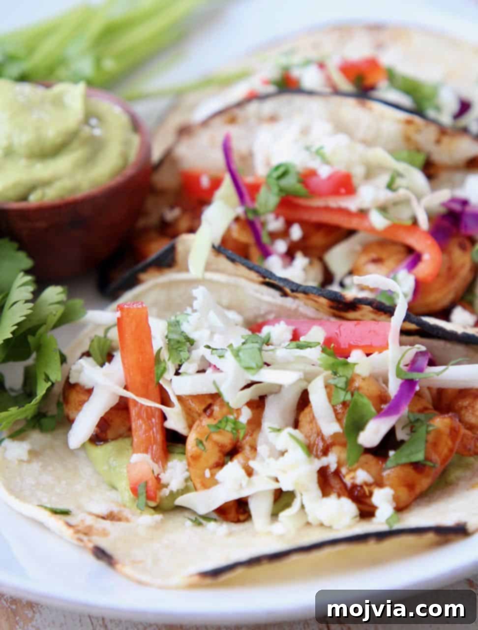 Zesty Grilled Shrimp Tacos with Creamy Avocado Salsa 4 Tacos filled with shrimp, slaw and avocado salsa on white plate with fresh cilantro