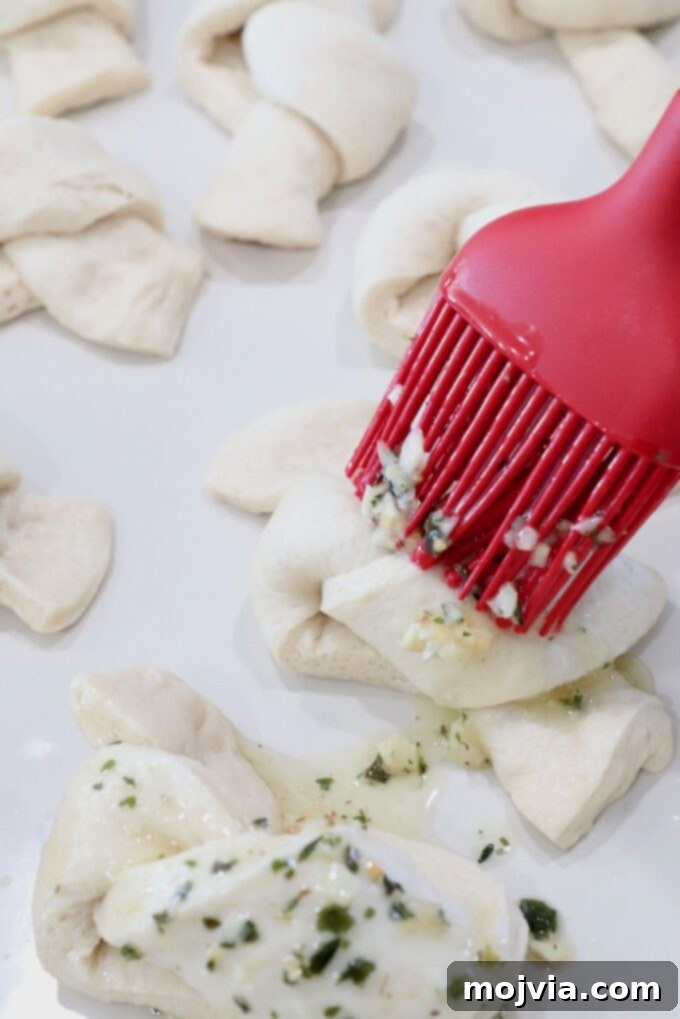 A pastry brush applying the seasoned garlic butter mixture onto uncooked garlic knots arranged on a baking sheet.
