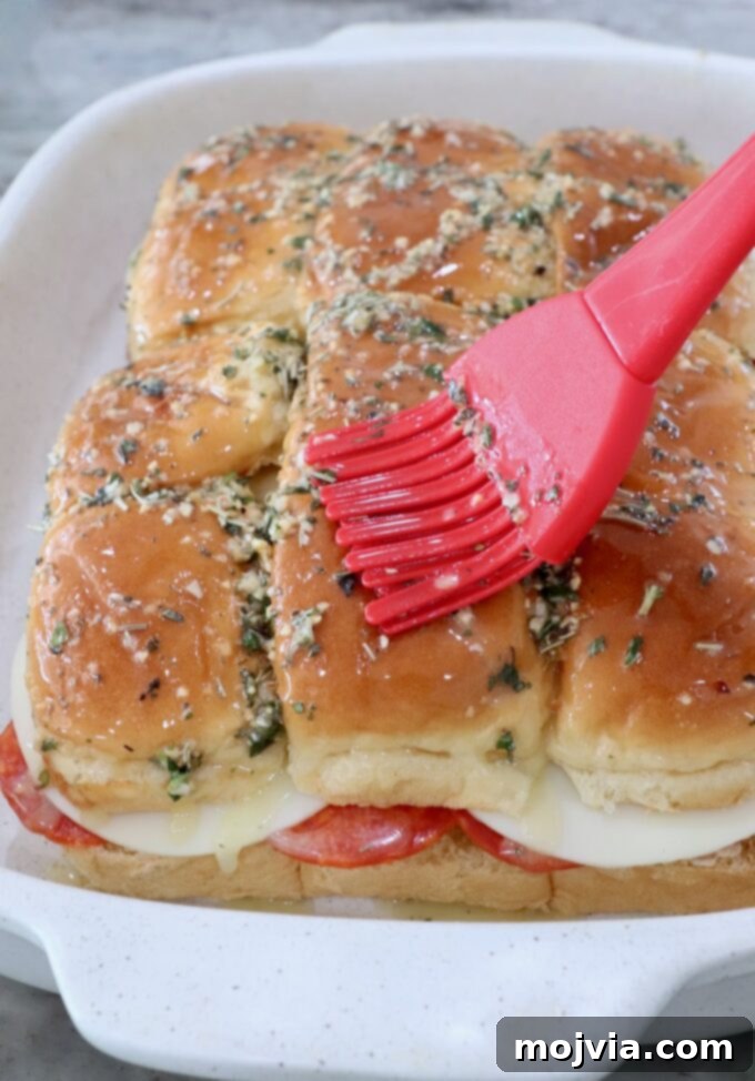 Italian sub sliders in a casserole dish, with the herb butter mixture generously brushed over the top of the rolls, ready for baking.