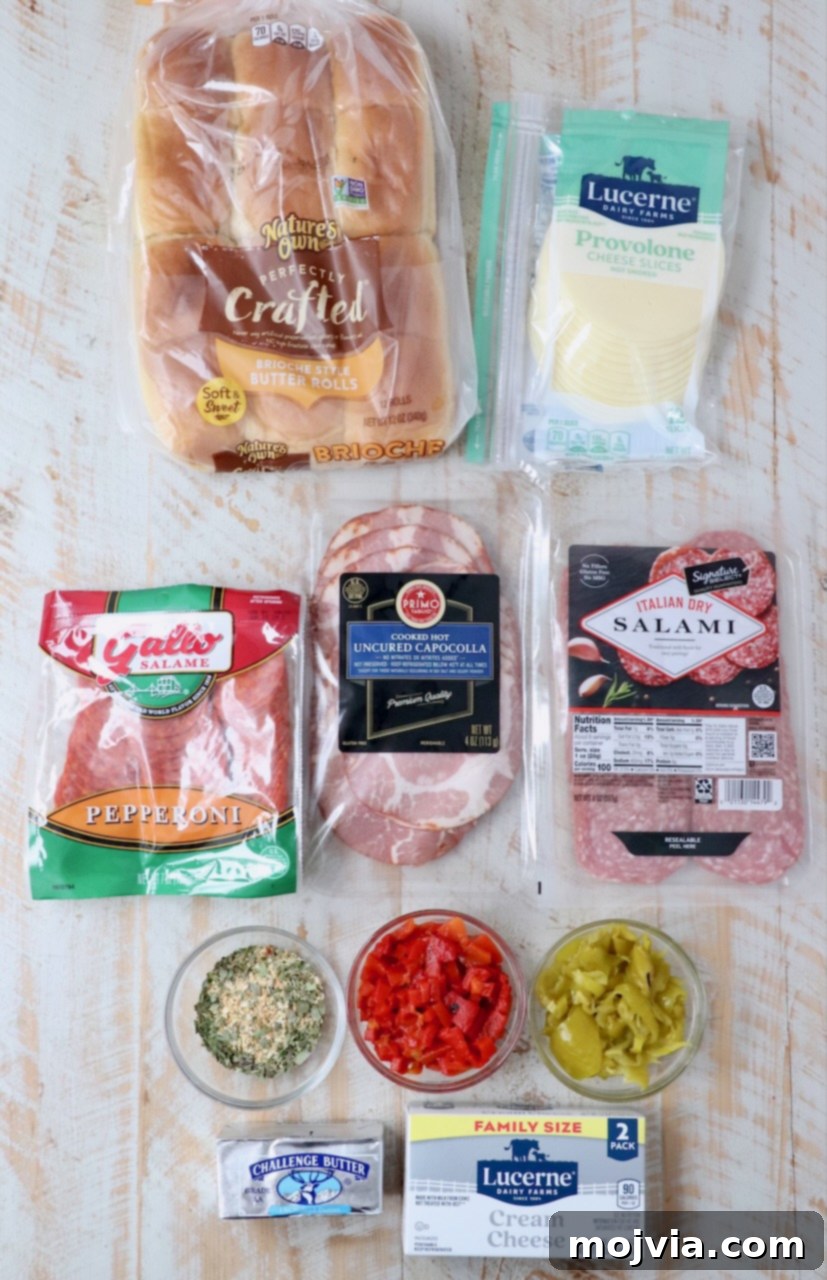 Ingredients for making Italian sub sliders laid out on a white wooden board, including rolls, deli meats, cheeses, and condiments.