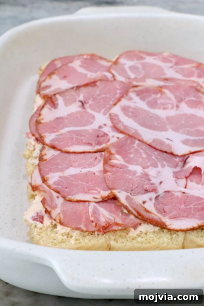 A layer of thinly sliced capocollo meat being added over the seasoned cream cheese on the bottom half of the rolls in the casserole dish.