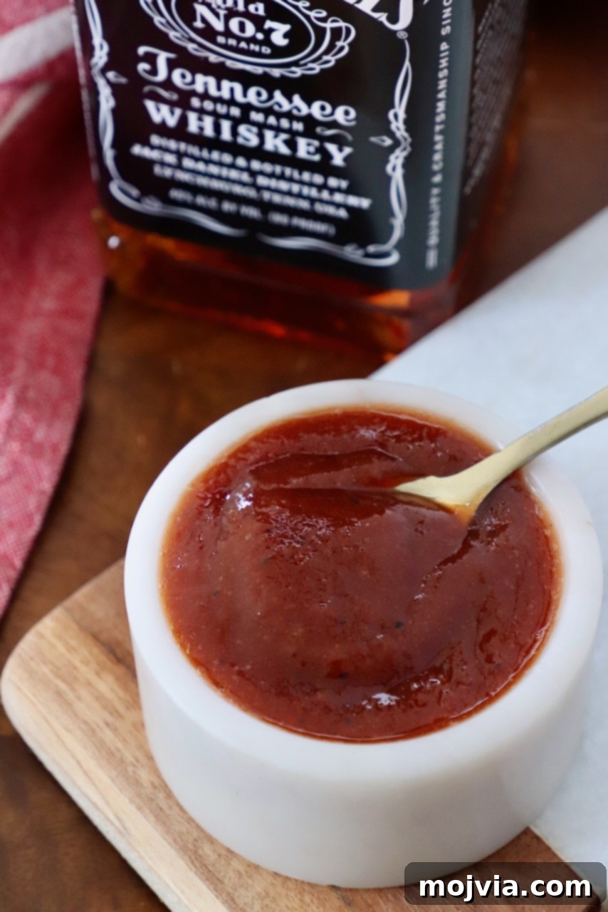 Jack Daniel's: The Taste of Tennessee BBQ 2 A small bowl of glistening homemade Jack Daniel's BBQ sauce with a spoon, accompanied by a bottle of Jack Daniel's whiskey, against a rustic background.