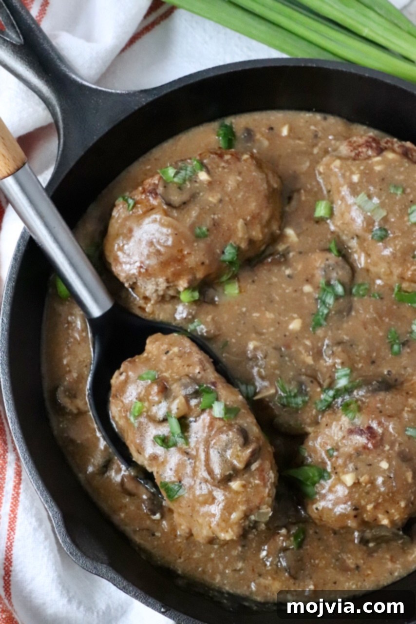 A close-up of Salisbury Steak in a skillet, richly covered with mushroom gravy, with a serving spoon ready to scoop.
