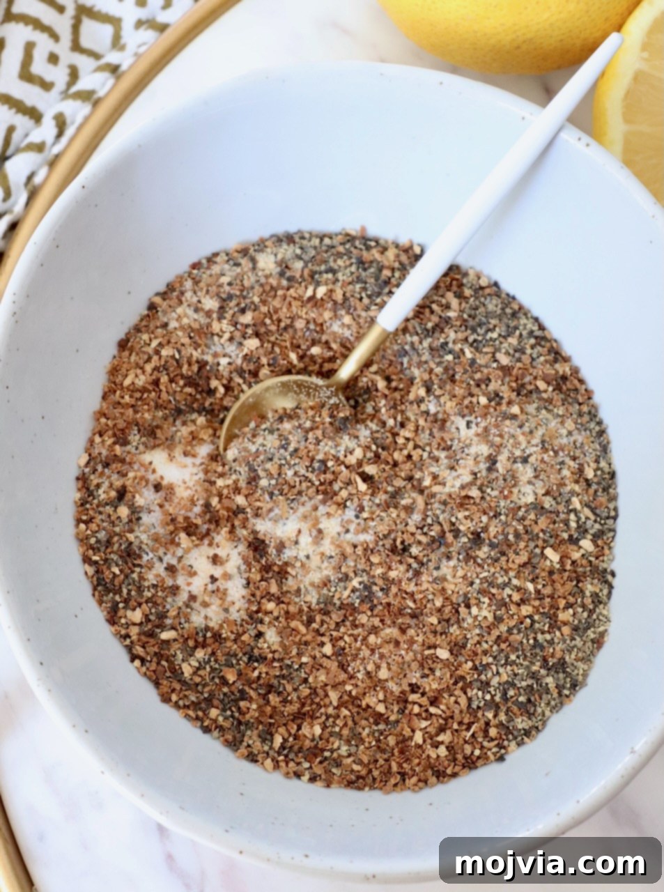 Zesty Lemon Pepper Blend 2 A close-up shot of various spices expertly mixed together in a small bowl, accompanied by a tiny spoon, ready for creating homemade lemon pepper seasoning.