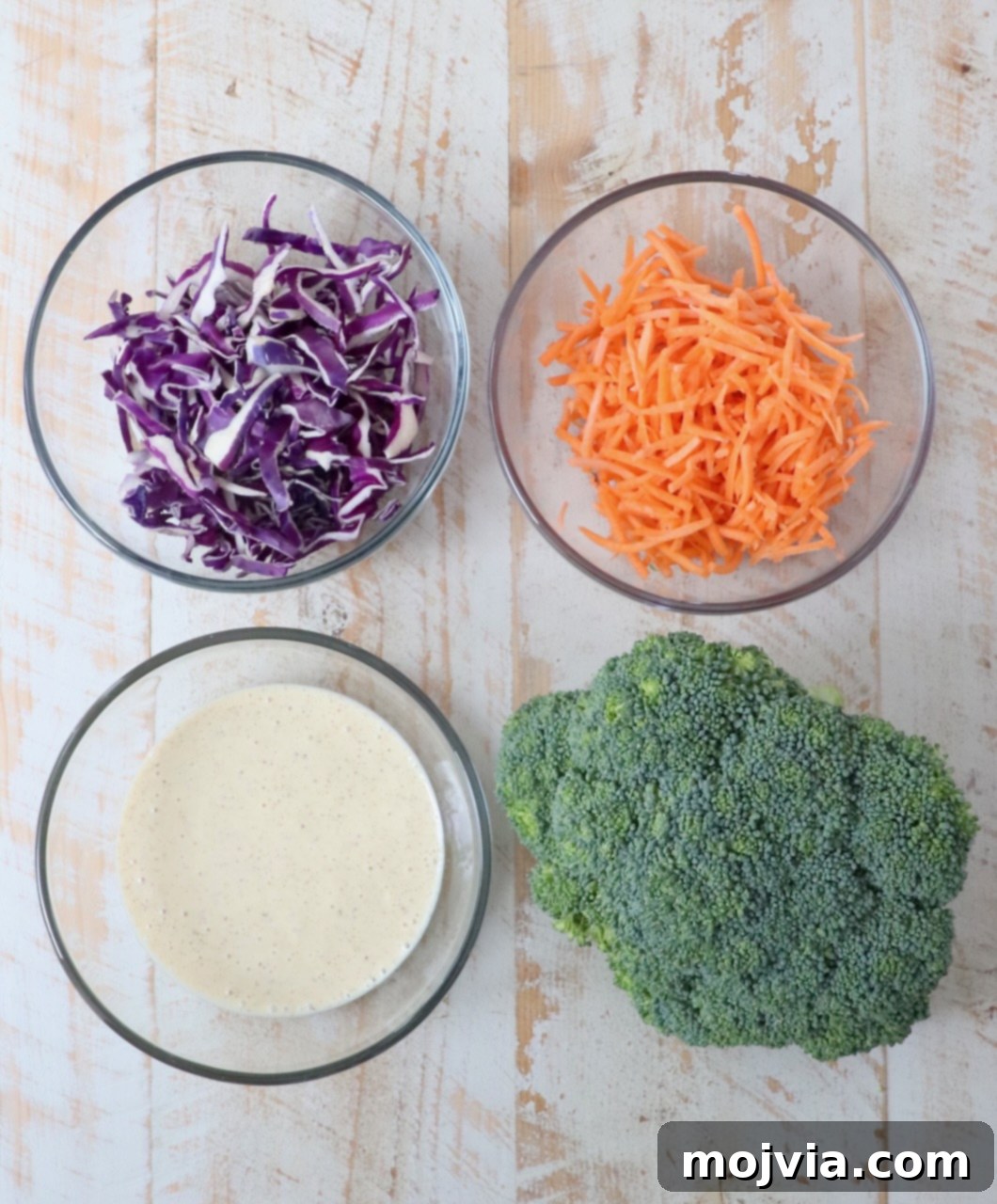 Vibrant Broccoli Slaw 3 Fresh ingredients for homemade Broccoli Slaw laid out on a white wooden board, including a head of broccoli, shredded carrots, and red cabbage.