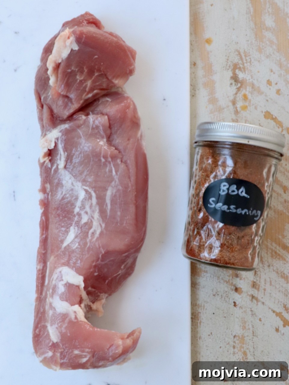 Fire-Kissed Pork Tenderloin 3 Raw pork tenderloin next to a small bowl of homemade BBQ seasoning on a rustic white wooden board, ready for preparation.