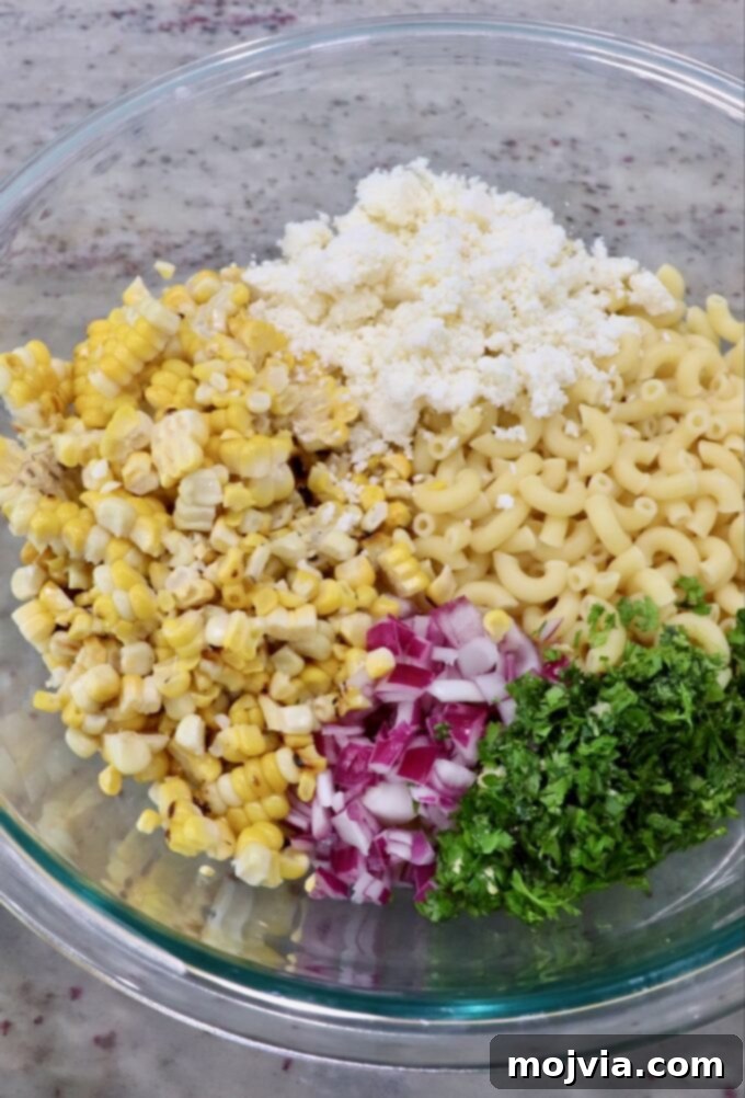 Cooked macaroni pasta in a large glass mixing bowl, with diced red onions, fresh cilantro, charred corn kernels, and crumbled cotija cheese added.