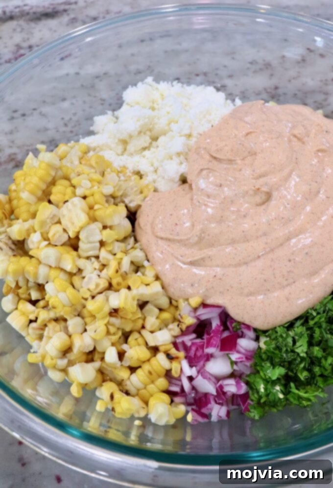 The creamy chili lime dressing being poured over the pasta, corn, onion, cilantro, and cotija cheese in the large glass bowl.
