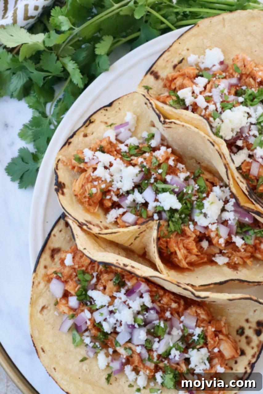 Smoky Chipotle Chicken Tinga 8 three tacos on plate filled with chicken tinga, cotija cheese, cilantro and onions