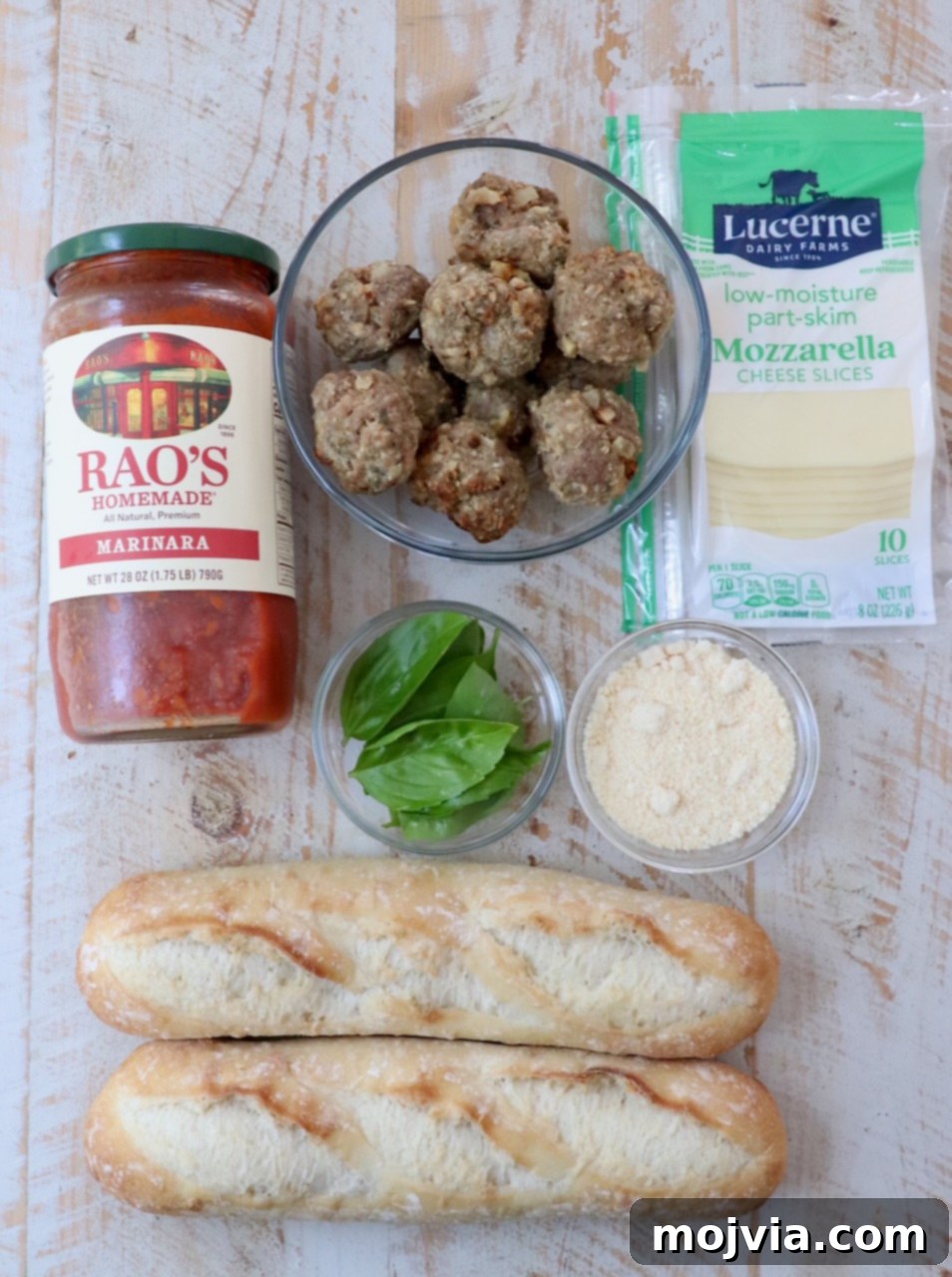 A selection of ingredients for making meatball subs laid out on a white wooden board, including meatballs, baguette, marinara, and cheeses.