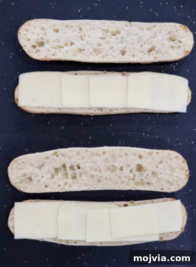 Two demi baguettes sliced lengthwise and topped with mozzarella cheese slices on a cutting board.