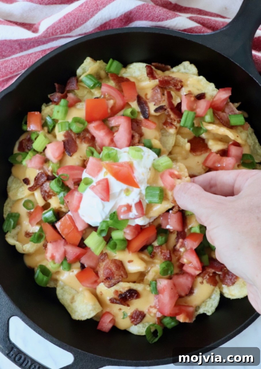 Emerald Potato Nachos 7 A hand reaches into a cast iron skillet of Irish nachos, dipping a chip into the creamy sour cream center.