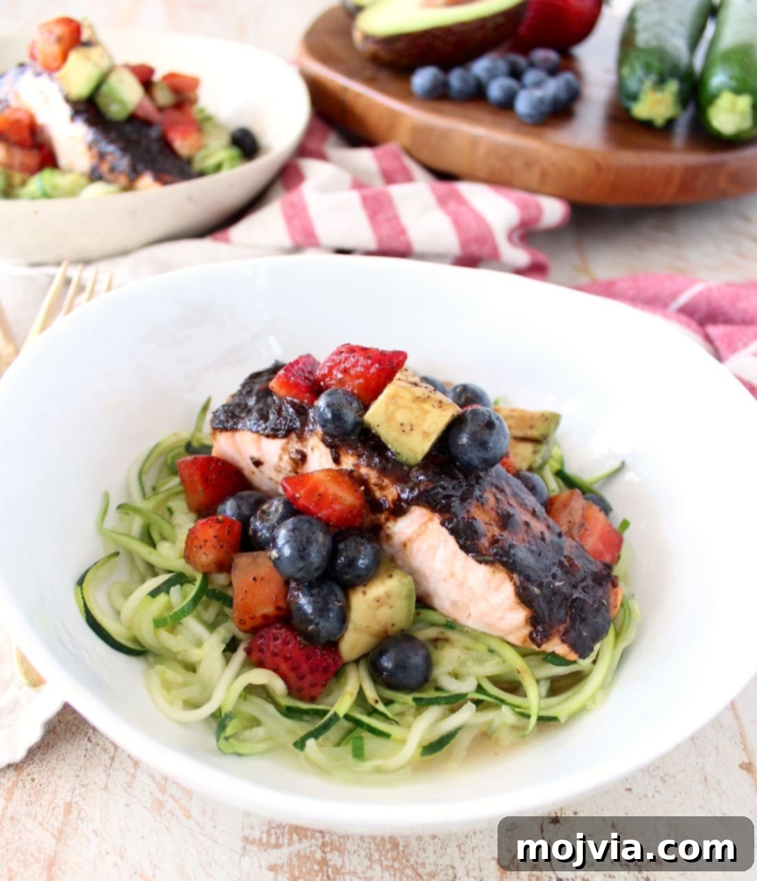 Balsamic Salmon Zoodle Bowls Roasted balsamic salmon is served over zucchini noodles topped with a berry avocado salsa in a white bowl.