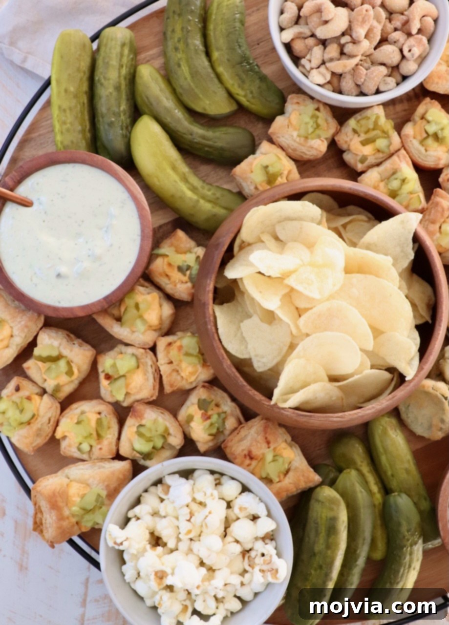 A large round charcuterie board overflowing with various pickle-flavored snacks, including chips, dips, popcorn, and whole pickles, set against a rustic background.